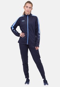 Marine Blau Trainingsanzug mit hellblauen Akzenten, bestehend aus einer Jacke mit Reißverschluss und Hose. Hergestellt aus einem glatten, atmungsaktiven Stoff, mit Logo- und Knopfdetails.