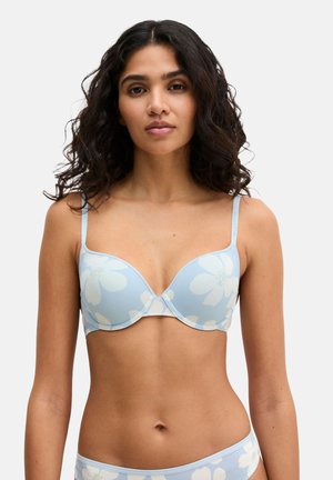 Femme portant un soutien-gorge bleu clair et une culotte assortie avec un grand motif floral blanc, debout devant un fond blanc uni.