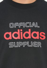 Melns kokvilnas T-krekls ar uzrakstu "OFFICIAL adidas SUPPLIER" izteiksmīgā baltā un sarkanā krāsā, apaļa kakla izgriezuma un īsām piedurknēm.