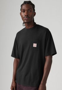 Black cotton t-shirt with a crew neck and short sleeves, featuring a small left chest pocket with a red label. Smooth texture and relaxed fit.