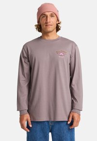 Długorękawowa bluzka w kolorze mauve z prążkowanymi mankietami i małym różowym logo na klatce piersiowej. Noszona z niebieskimi dżinsami i jasnoróżową czapką.