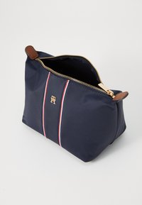Marineblå nylon kosmetikkveske med gullglidelås, brune lærdetaljer og røde og hvite striper på fronten. Firkantet form.