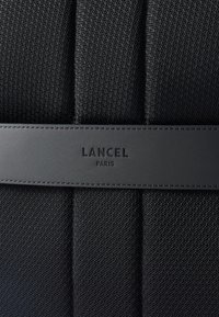 Fekete textil táska, amely strukturált dizájnnal rendelkezik, sima, dombornyomott bőr sávval. A középen hangsúlyosan szerepel a "LANCEL PARIS" felirat.
