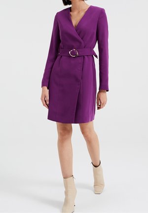Paarse wikkeljurk met lange mouwen, een taille met een riem en een ronde gesp, en een rechte rok. Gecombineerd met beige enkelhoge laarzen.
