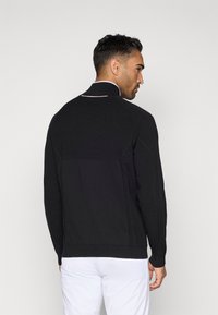 Μαύρο αθλητικό μπουφάν με ψηλό γιακά, υφή υφάσματος και λευκή περιφέρεια στον γιακά. Ορατοί ραφές και εφαρμοστή σιλουέτα.