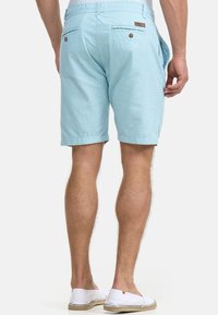 Mann trägt hellblaue knielange Shorts und weiße Slipper, steht mit seitlich hängenden Händen, Rückansicht vor weißem Hintergrund.