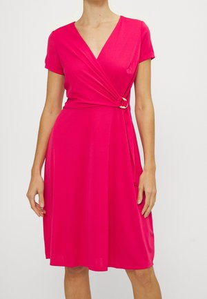 Femme portant une robe portefeuille rose vif mi-genou avec des manches courtes et un détail de bague dorée à la taille, debout devant un fond uni.