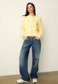 Cardigan jaune et duveteux avec un col rond et une fermeture à boutons, associé à un jean bleu clair à jambes larges et des baskets blanches.