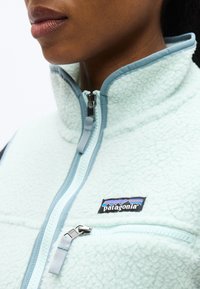 Κοντινό πλάνο ενός ατόμου που φοράει ένα γαλάζιο φλις μπουφάν με ψηλό γιακά, μπροστινό φερμουάρ και εμφανές λογότυπο Patagonia στο στήθος.