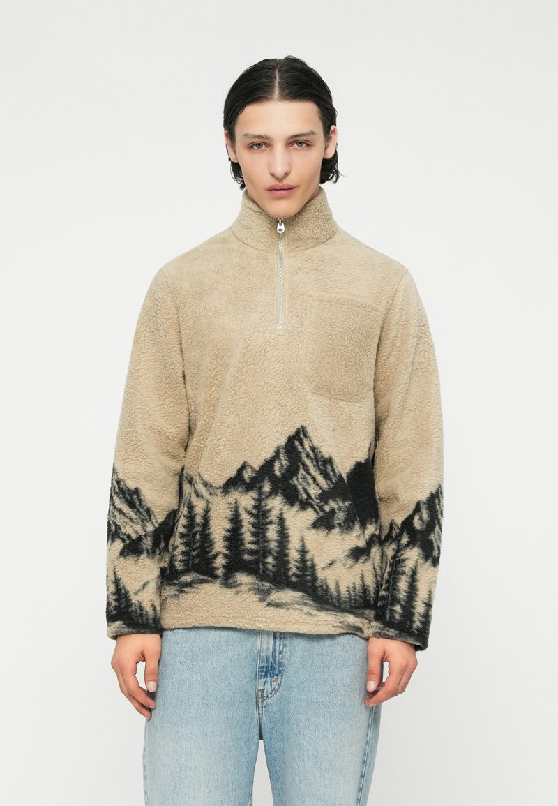 Beige Fleece-Pullover mit hohem Kragen, der am Saum ein schwarzes Berg- und Walddesign aufweist. Verfügt über eine Brusttasche und einen Halb-Reißverschluss.