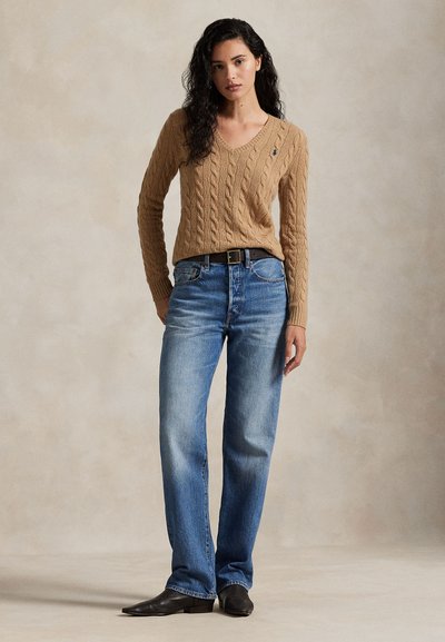 Žena s dlhými kučeravými vlasmi, oblečená v karamelovom svetríku s vzorom a výstrihom do V, modré džínsy, čierny opasok a čierne špicaté topánky, stojaca na neutrálnom pozadí.