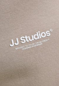 Logo kujundus, millel on "JJ Studios" valges tekstis tekstuurses, heledas beežis kangas, all olevate väiksemas kirjas tekstidega.
