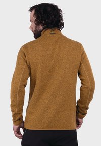 Braune Fleecejacke mit hohem Kragen, strukturiertem Stoff, kontrastierenden Nähten und kleinem Logo am Nacken.