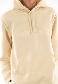 Felpa color crema con tasca frontale, cappuccio con cordino e logo "Timberland" in rilievo. Tessuto morbido e design minimale.