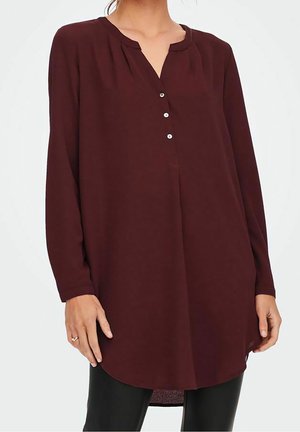 Frau trägt eine dunkel burgunderrote Tunika-Bluse mit langen Ärmeln, einer teilweise geknöpften Leiste und abgerundetem Saum, kombiniert mit einer schwarzen Hose.