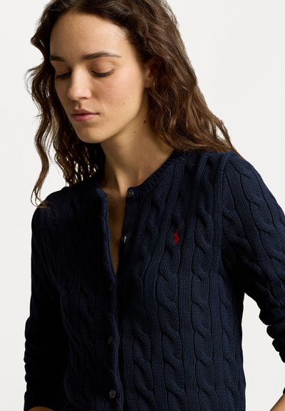 Navy blue cable-knit cardigan with a button-up front, ribbed collar and cuffs, and a small red logo on the left chest area.