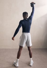 Juodas sportinis marškinėlis, pilkos šortai su šoninėmis juostelėmis, baltos sportbačiai. Asmuo pakelia kettlebell virš galvos minimalistinėje aplinkoje.