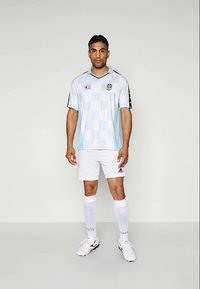 Šviesiai mėlynas futbolo marškinėliai trumpomis rankovėmis su šaškių raštu, juodomis detalėmis ir logotipu; derinami su baltais šortais ir kojinėmis bei juodomis sportbačiais.