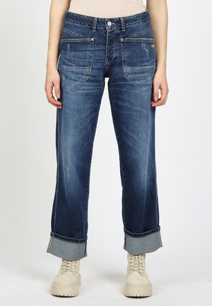 Dunkelblaue, weit geschnittene Jeans mit aufgeschlagenen Bündchen und großen Fronttaschen, kombiniert mit beigen Schnürplattformstiefeln.