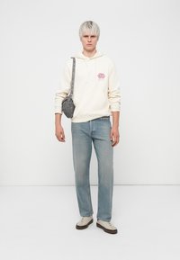 Light beige hoodie with red text, blue denim jeans, gray crossbody bag with silver accents, and cream shoes with brown soles.