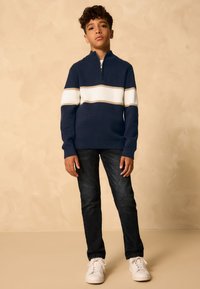 Laivastonsininen pusero, jossa valkoiset ja beigen raidat, edessä vetoketju ja resorit hihansuissa, yhdistetty tummiin farkkuihin ja valkoisiin lenkkareihin. Yksinkertainen muotoilu.