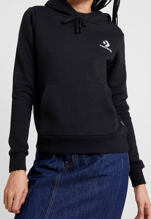 Personne portant un sweat à capuche noir Converse avec une poche avant et une jupe en jean, montrant le haut du corps du menton jusqu'aux hanches.