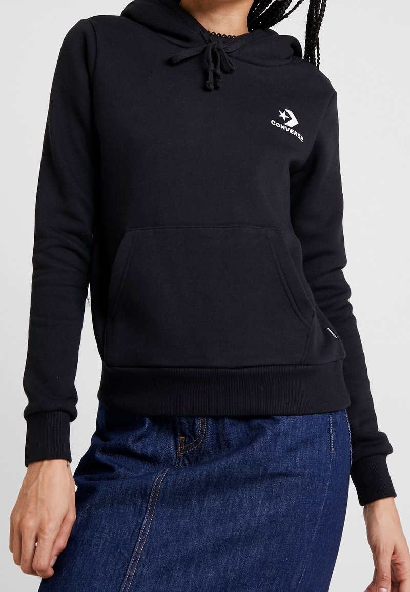 Personne portant un sweat à capuche noir Converse avec une poche avant et une jupe en jean, montrant le haut du corps du menton jusqu'aux hanches.