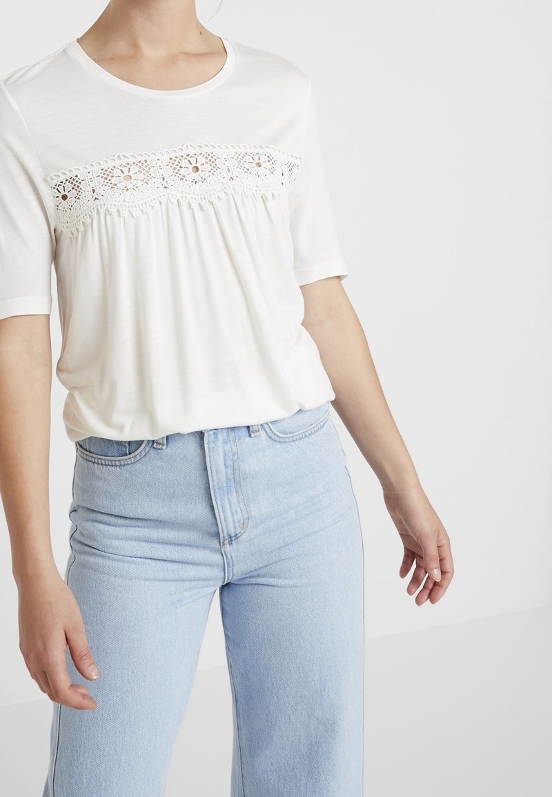 Vit kortärmad topp med spetsdetaljer över bröstet, kombinerad med ljusblå högmidjade jeans. Mjuk, texturerad tyg.