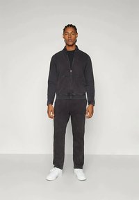 Chaqueta negra con cremallera y bolsillos frontales, junto con pantalones a juego, ambos hechos de tela suave; combinados con zapatillas blancas, creando un look casual.