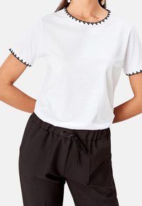 Maglietta bianca con ricamo triangolare nero lungo il collo e le maniche, abbinata a pantaloni neri con coulisse e fronte plissettato.