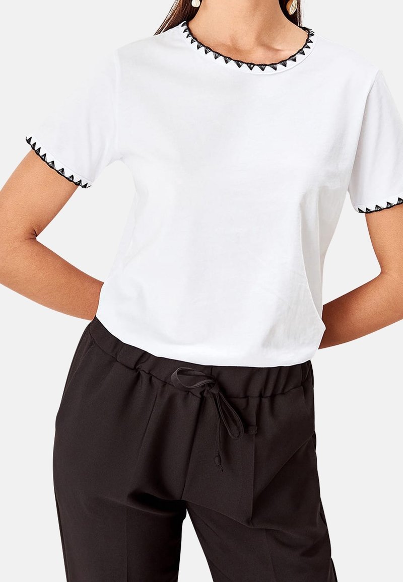 Maglietta bianca con ricamo triangolare nero lungo il collo e le maniche, abbinata a pantaloni neri con coulisse e fronte plissettato.