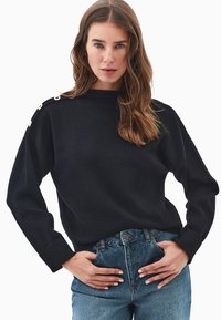 Schwarzer Strickpullover mit hohem Kragen und überschnittenen Schultern. Besitzt Knopfakzente an der linken Schulter. Kombiniert mit hochgeschnittenen Denim-Jeans.