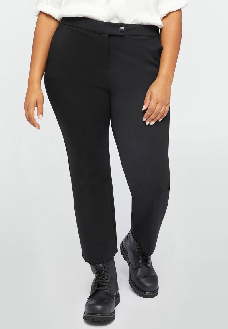 Pantaloni negri cu un design ajustat, având închidere cu nasture și buzunare. Purtați cu bocanci negri cu șireturi. Textură netedă, croială dreaptă.