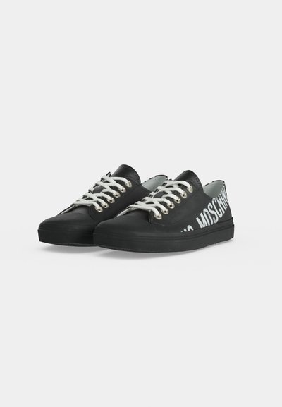 Baskets basses noires avec lacets blancs et grand marquage blanc Moschino sur les côtés extérieurs, présentées sur fond blanc.
