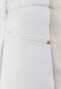 Tela de mezclilla blanca con una textura suave, que presenta un bolsillo lateral y detalles de hardware en tono cobre. El diseño resalta las costuras en contraste.