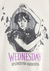 Vit t-shirt med en grafisk design av en kvinna med mörkt hår, iklädd en volangkrage, omgiven av flugande svarta fåglar. Texten nedanför lyder "ONSDAG BESTÄMD FÖR MÖRKER."
