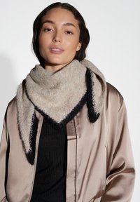 Sciarpa triangolare beige con accenti neri, realizzata in tessuto morbido e testurizzato, drappeggiata intorno al collo, abbinata a una giacca di satin.