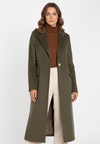 Olivengroene wollen jas met een geremde reverskraag, enkele knoopsluiting en leopardprint voering; gedragen over een bruine coltrui en crèmekleurige broek.