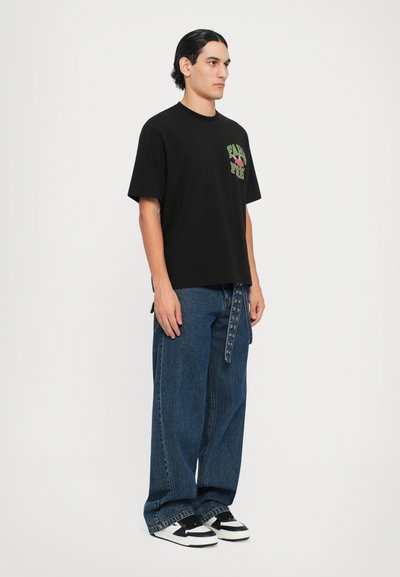 Black cotton t-shirt with green and pink graphic, paired with wide-leg blue denim jeans and black and white sneakers with laces.