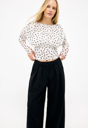 Femme blonde portant un chemisier blanc à manches longues à pois et un pantalon noir ample taille haute, debout les mains près des poches.