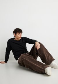 Jeune homme assis par terre portant une chemise noire à manches longues, un pantalon marron à jambes larges et des baskets blanches, sur un fond blanc uni.