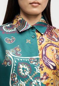 Femme portant une chemise boutonnée avec des panneaux turquoise et jaune moutarde ornés de motifs floraux et paisley complexes.