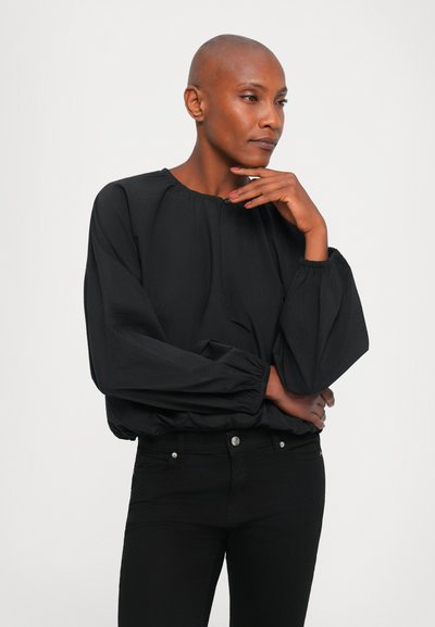 Blusa nera leggera con scollo rotondo e polsini elasticizzati. Presenta tessuto arricciato e una vestibilità leggermente morbida. Abbinata a jeans neri.
