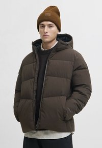 Jeune homme portant une veste matelassée marron avec les mains dans les poches et un bonnet en tricot marron sur fond clair.