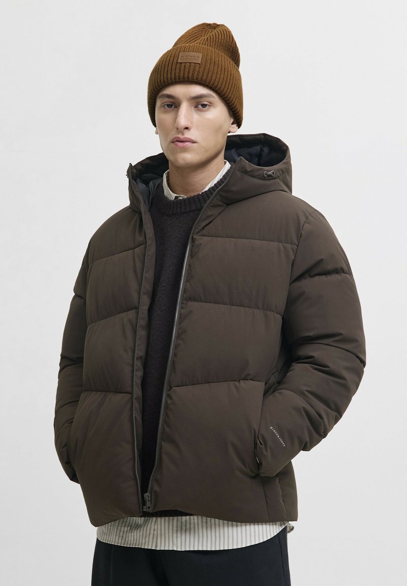 Jeune homme portant une veste matelassée marron avec les mains dans les poches et un bonnet en tricot marron sur fond clair.