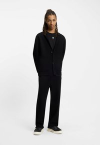 Musta bleiseri, jossa on lovettu kaulus ja kaksi etutaskua, yhdistettynä vastaaviin mustiin housuihin ja mustiin lenkkarisiin, joissa on vaaleat pohjat.