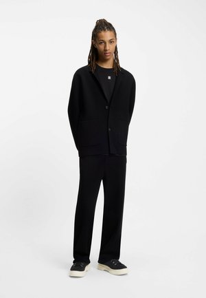 Musta bleiseri, jossa on lovettu kaulus ja kaksi etutaskua, yhdistettynä vastaaviin mustiin housuihin ja mustiin lenkkarisiin, joissa on vaaleat pohjat.