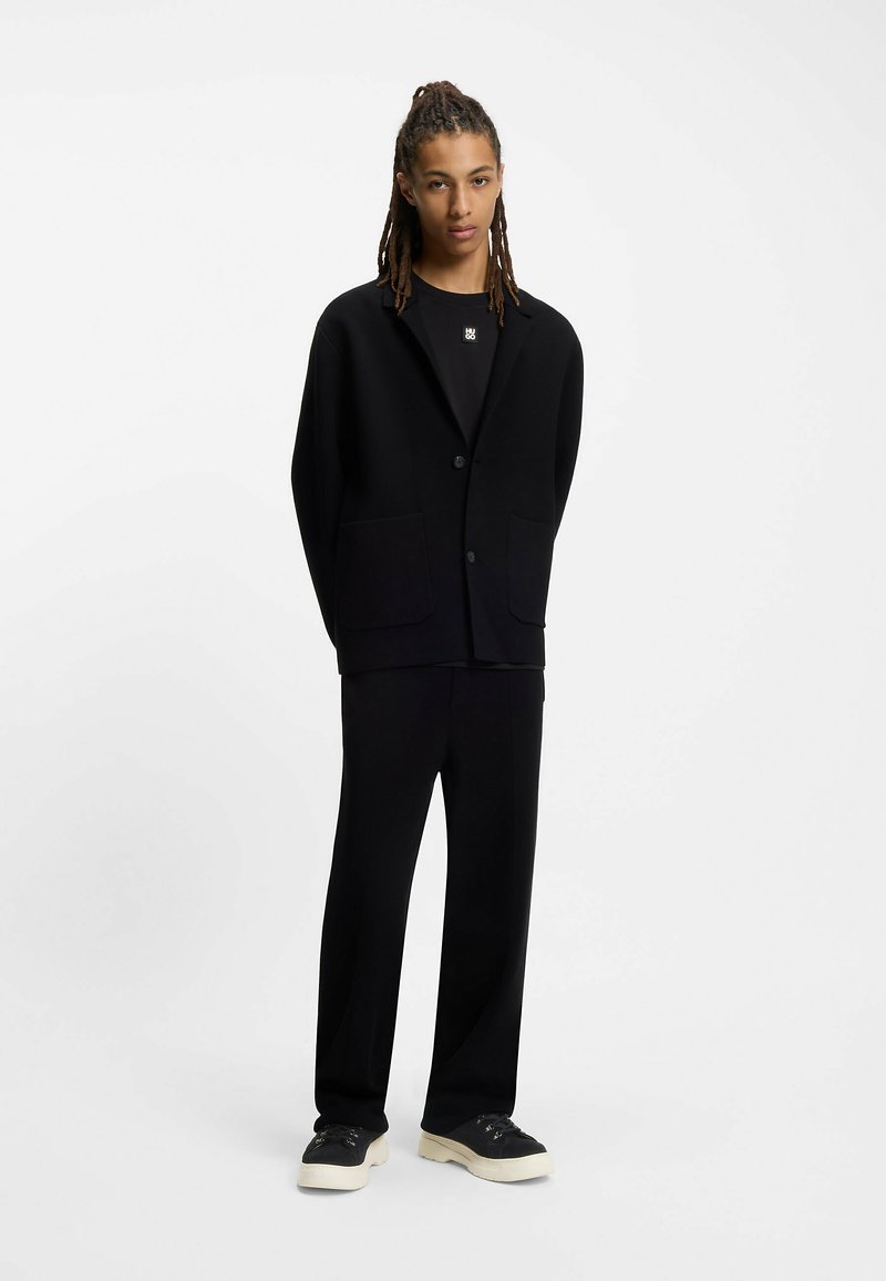 Blazer nero con colletto a risvolto e due tasche frontali, abbinato a pantaloni neri coordinati e sneakers nere con suola crema.