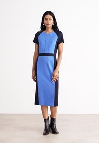 Μπλε και ναυτικές αποχρώσεις στρατηγικά τοποθετημένες σε φόρεμα με κοντά μανίκια, εφαρμοστή σιλουέτα και μήκος μέχρι το γόνατο. Διαθέτει στρογγυλή λαιμόκοψη και διακριτές ραφές.