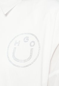 Tricou polo alb cu guler, având un logo circular cu aplicare de pietre strălucitoare argintii pe partea stângă. Textură netedă de bumbac.